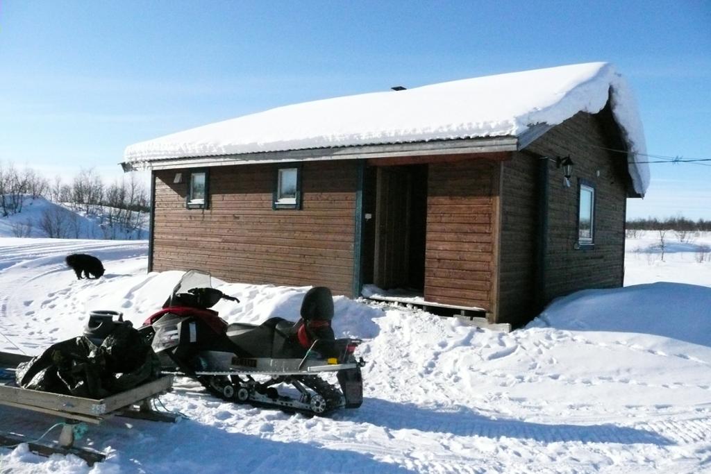 Liten brun hytte med snøskuter foran. Foto.