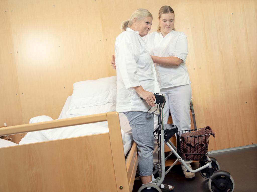 Kvinnelig pasient får hjelp ut av senga og bort til rullatoren sin. Foto.