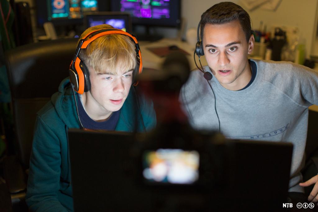 Two teenage boys with headsets look at a computer screen. Behind them are more screens. Photo.