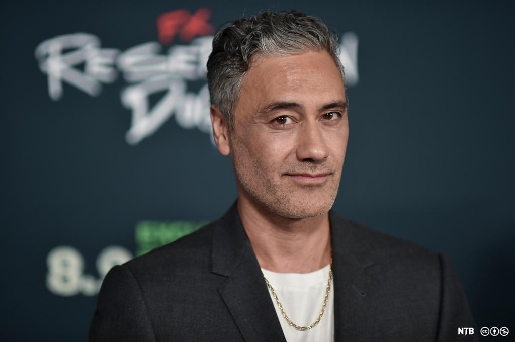 Photo: Portrait of Taika Waititi in front of a black wall where we can see part of the title 'Reservation Dogs'. He is wearing a black suit jacket, white T-shirt, and a gold necklace. 
