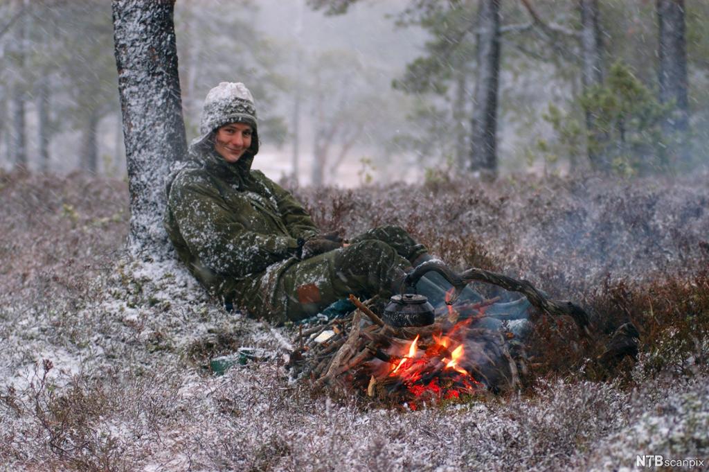En person sitter ved et bål i skogen. Det er snø i lufta. Foto.