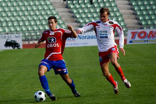 To fotballspelarar frå kvart sitt lag spring etter ein ball framfor tomme tribunar. Foto.