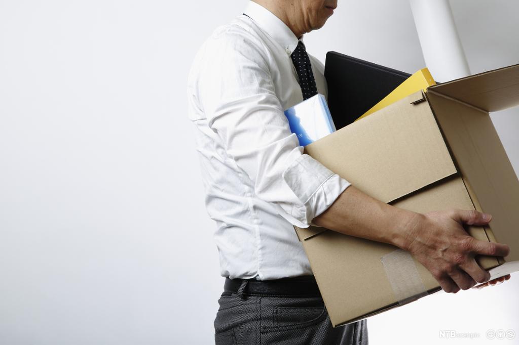 Mann med skjorte, dressbukse og slips bærer en pappeske med kontorsaker. Foto.