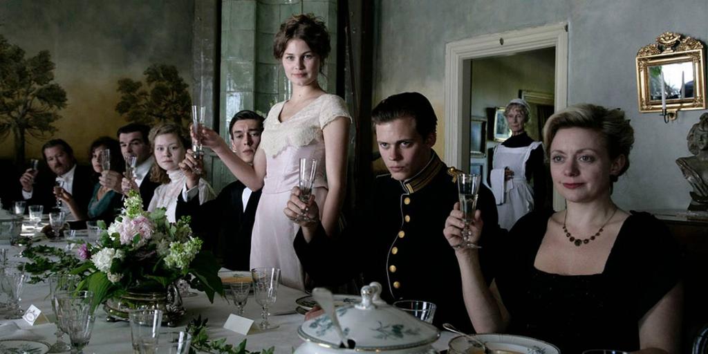 Stillbilde fra filmen Victoria. Pent kledde personer sitter ved et dekket langbord og løfter glasset. En ung kvinne ved midten av bordet står oppreist, og de andre sitter. 