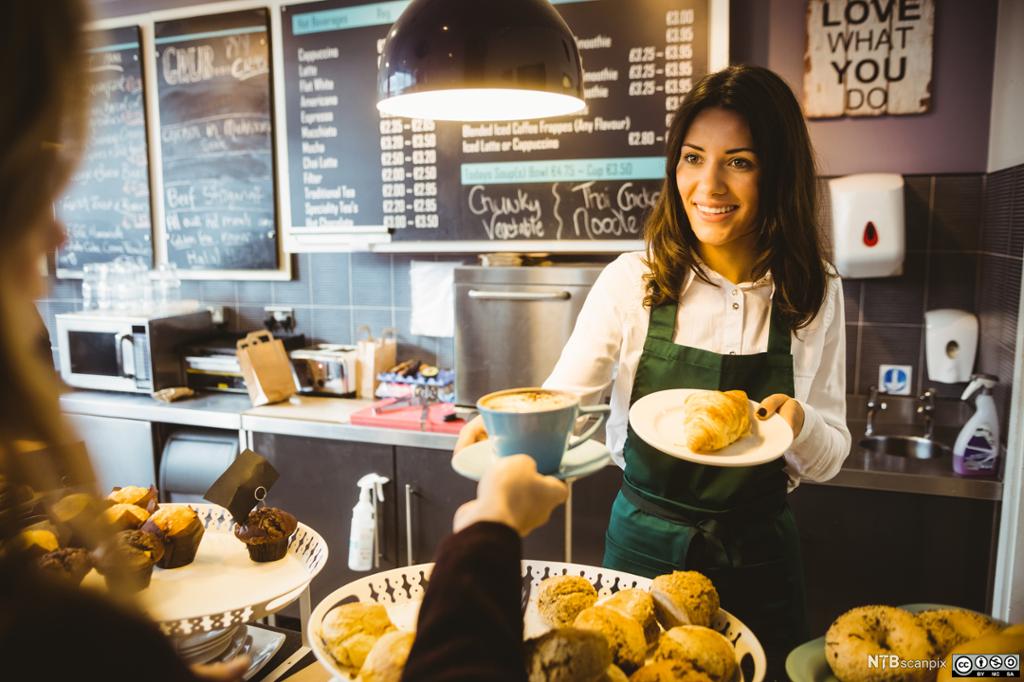 Bilde av kvinne i en kafé som rekker en kaffe og en croissant til en gjest. Foto. 