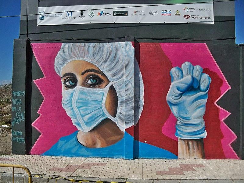 Photo: We see a sidewalk and a building. The building has a grafitti of a nurse in protective clothing. She is holding up a closed fist. The Spanish word VIDA (life) is written on the fingers of her  blue plastic glove. 