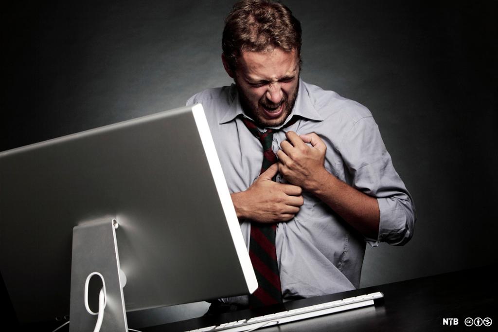 Mann sittende foran PC holder hendene foran brystet mens han skjærer grimaser. Foto. 