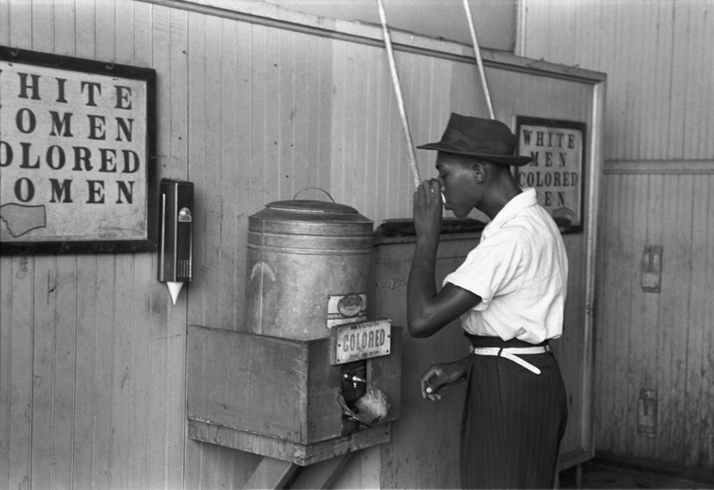 Photo: A Black man takes a drink from a drinking fountain labeled 'colored'. There are other signs with 'white' and 'colored' on the wall. 