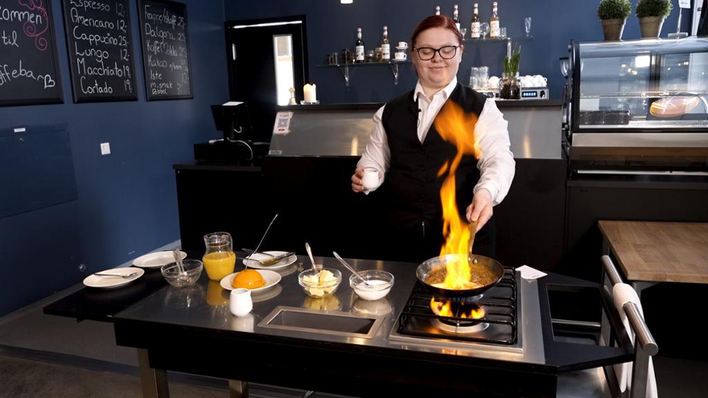 En ung kvinnelig servitør står ved en flamberingsvogn og flamberer en banan. Foto.