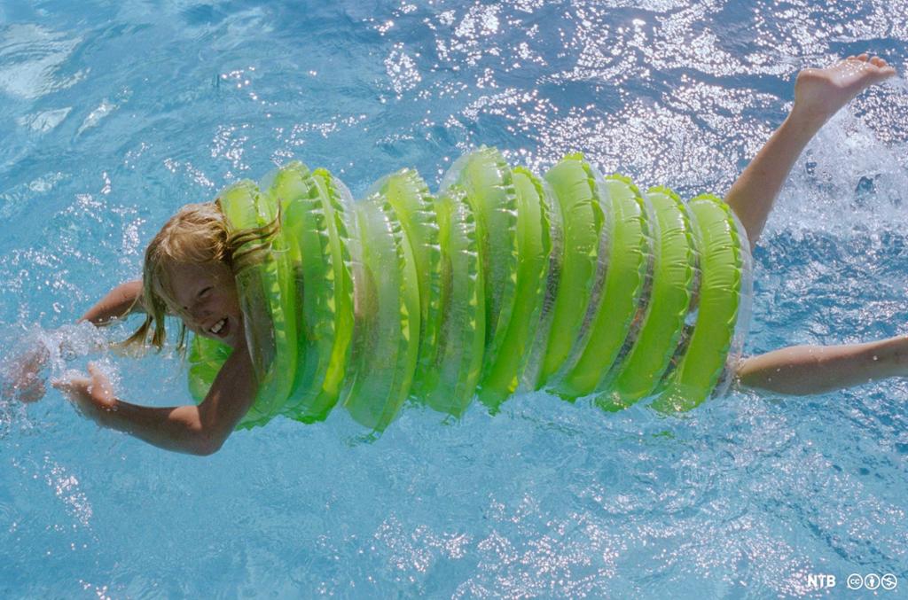 Et barn ler og svømmer med tolv baderinger rundt kroppen, bare armene, hodet og beina stikker ut. Foto.