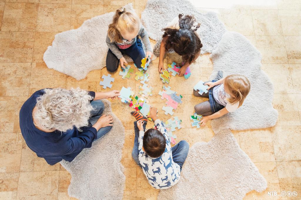 En voksen sitter på gulvet sammen med fire barn og legger et stort puslespill. Foto.