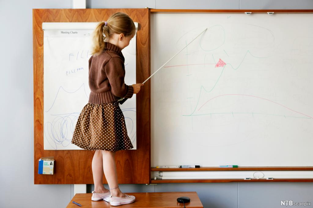 Skolejente leker at hun er lærer ved at hun peker med pekestokk på tavla. Hun står på en pult foran tavla. Foto.
