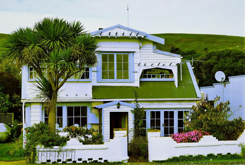 Photo: A white house with a green roof, blue details, and a white garden fence. It is surrounded by green nature. 
