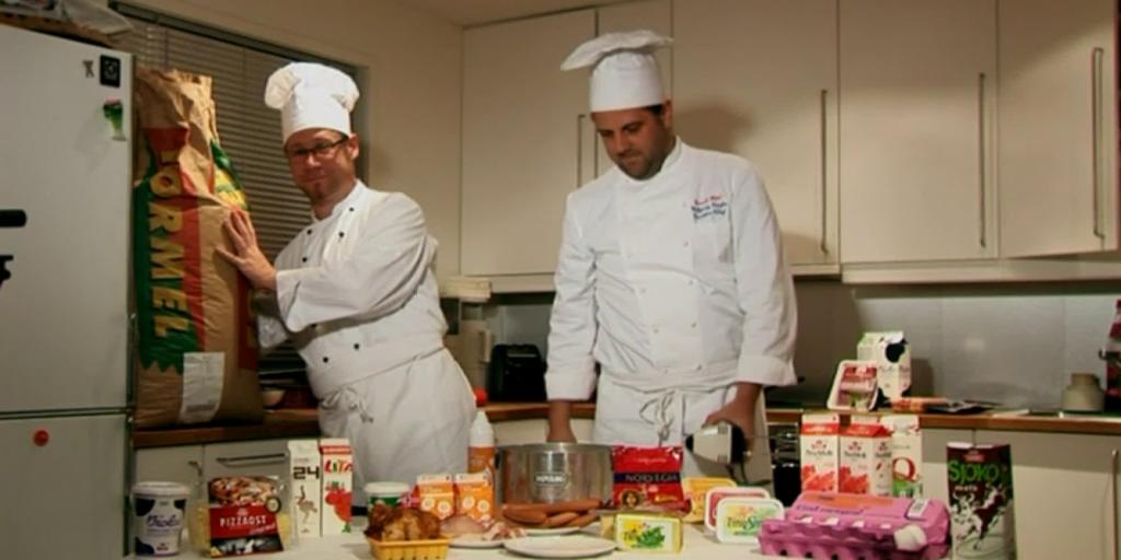 To menn i kokkeklær på et kjøkken med norske matvarer på kjøkkenbordet. Foto.
