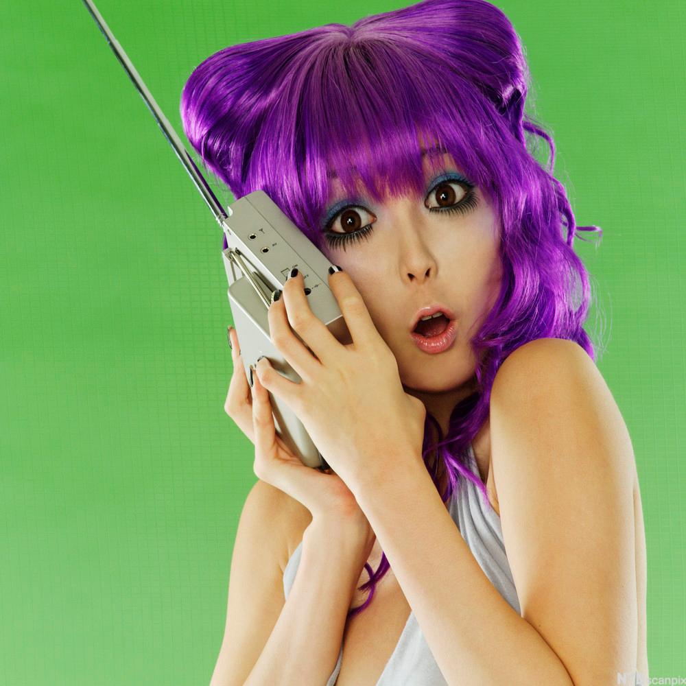 Ei jente med lilla hår og oppspila auge held ein radio tett inntil øyret og ser overraska ut. Foto.