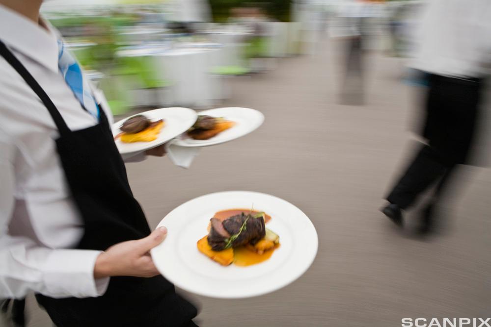 En servitør bærer tre tallerkener med mat ute i en restaurant. Foto.