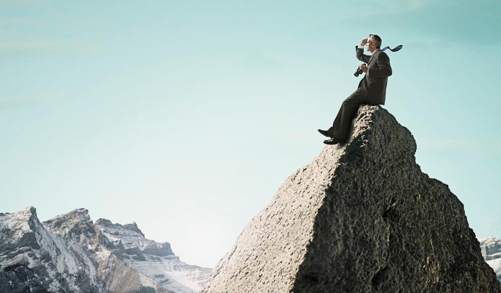 Ein dresskledd mann på toppen av eit fjell held ein kikkert og speidar utover. Foto. 