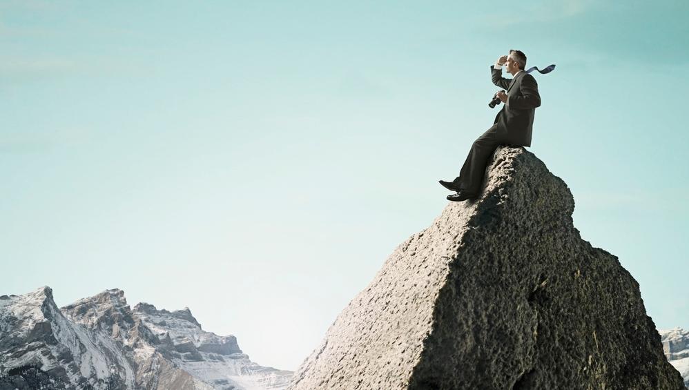 En mann i dress sitter på en fjelltopp og holder en kikkert. Foto.