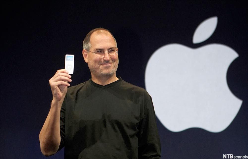 Steve Jobs viser fram en iPod musikkspiller. I bakgrunnen er Apple-logoen synlig. Foto.