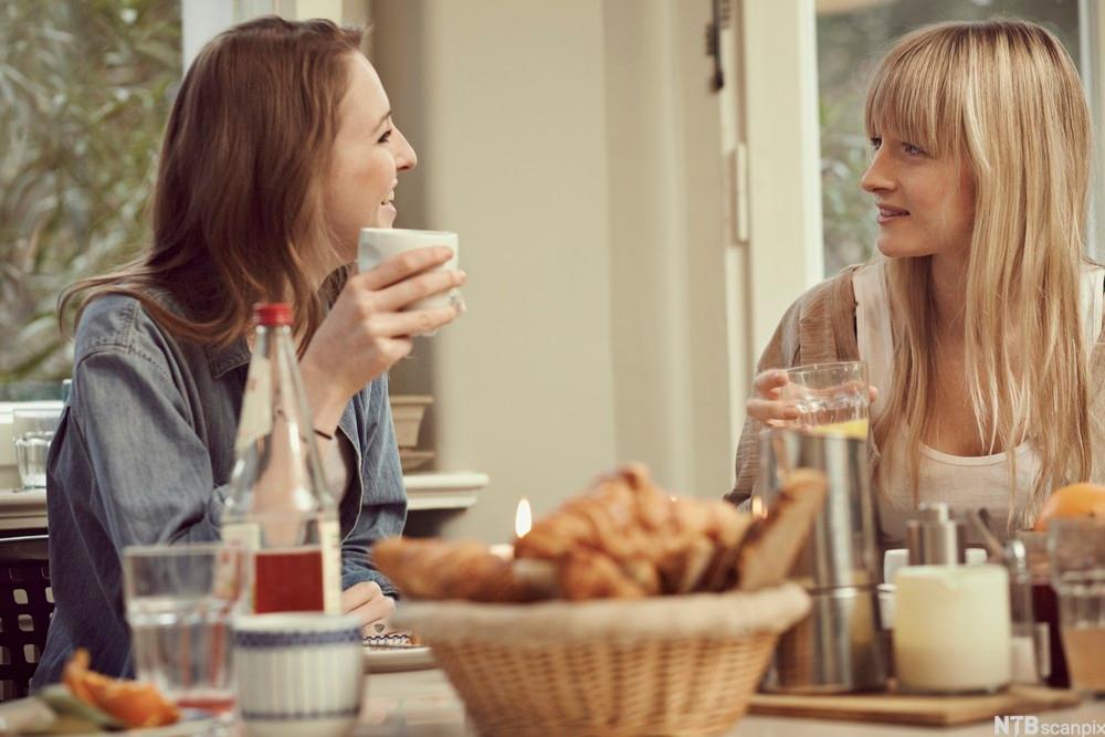 To kvinner sitter ved et bord dekket med mat og drikke. Kvinnene ser på hverandre og smiler. Foto.