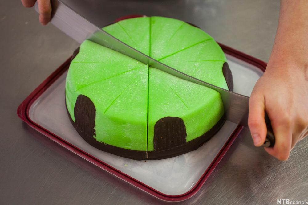 Ei grøn marsipankake blir delt med tohands kniv. Foto.