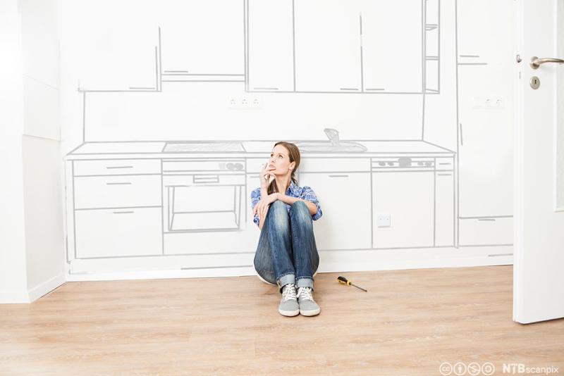 Kvinne sitter på gulvet foran et tegnet kjøkken og ser drømmende ut i lufta. Illustrasjon.