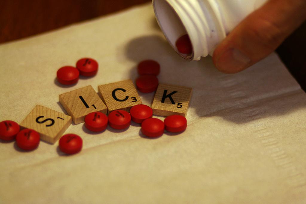 Ei hånd heller ut piller fra en pilleboks. Blant pillene ligger det fire brikker fra brettspillet Scrabble: bokstavene S, I C, K. Foto. 