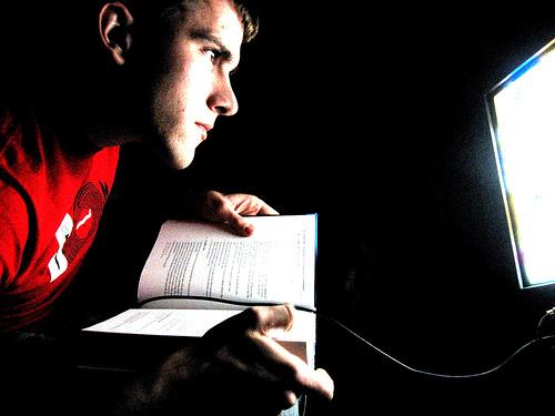 Gutt som studerer hardt. Han sitter med ei oppslått bok og ser inn i en dataskjerm. Foto. 