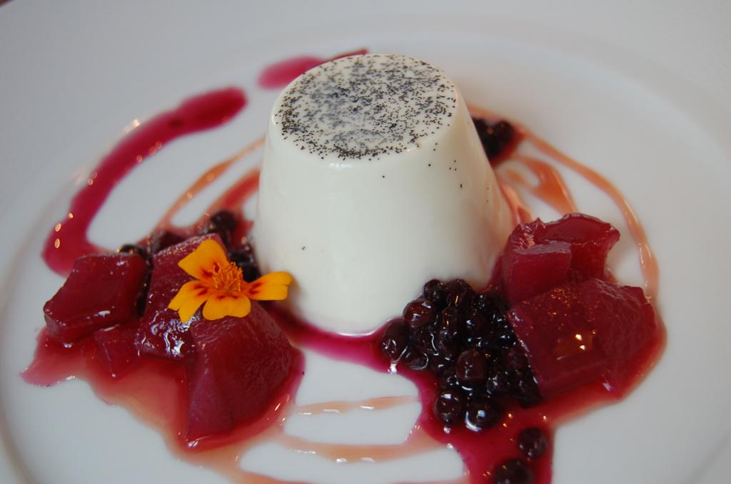 En hvelvet panna cotta på en tallerken, pyntet med rød saus, bærkompott og en gul blomst. Foto.