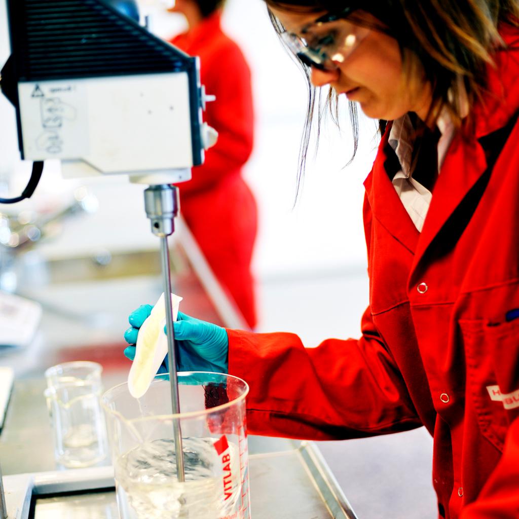 En laborant tilsetter salt i en løsning. Laboranten har rød labfrakk, plasthansker og vernebriller. Foto.