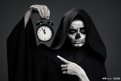 Mørk skikkelse som symboliserer døden holder klokke som viser fem på tolv. Foto.