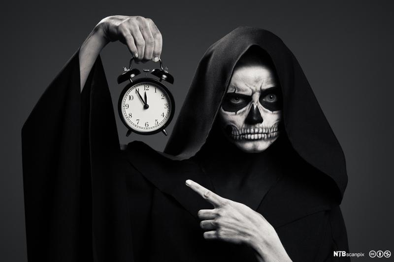 Mørk skikkelse som symboliserer døden holder klokke som viser fem på tolv. Foto.