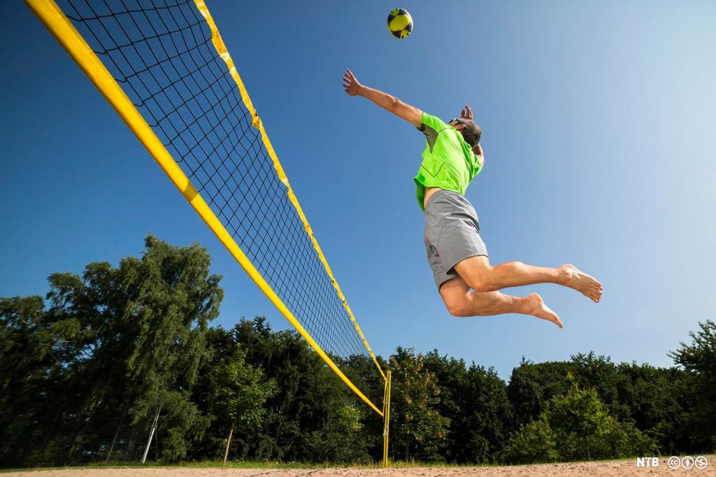 Mann i grå shorts og grønn trøye svever i lufta klar til å smashe en volleyball. Grønn løvskog og blå himmel i bakgrunnen. Foto.