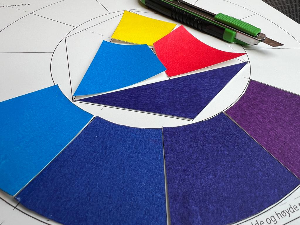 Tilskårne biter av papir malt i gult, rødt, blått og fiolett er plassert løst over en arbeidstegning av fargesirkelen. Foto.