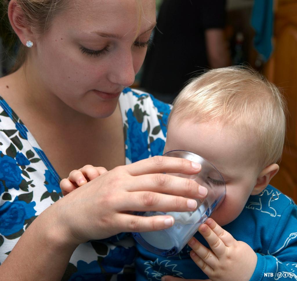En voksen kvinne har et lite barn på fanget. Hun holder et glass melk mot munnen til barnet. Barnet drikker og har også fingrene borti glasset. Glasset dekker nesten hele ansiktet til det lille barnet. Foto.