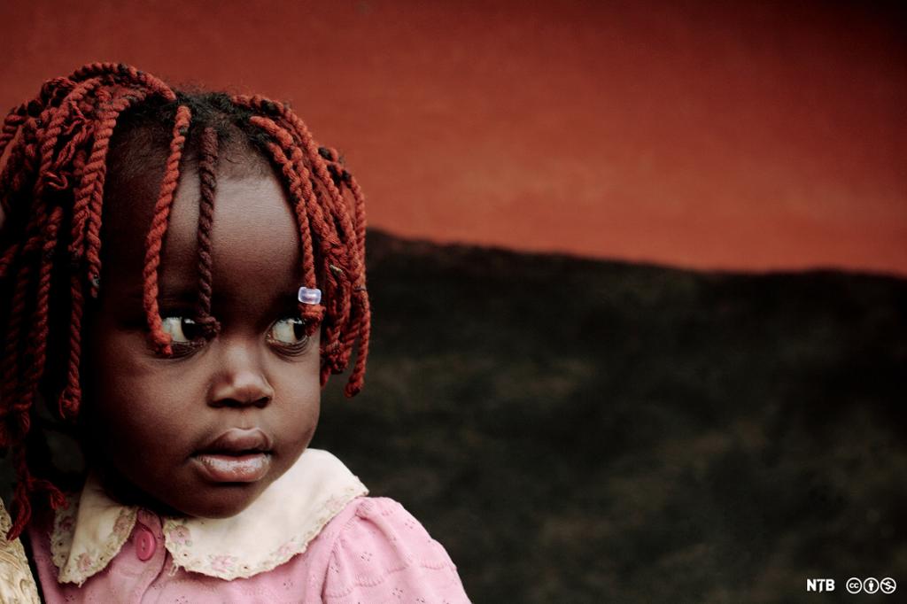 Nærbilde av en mørkhudet liten jente med rødfarget og flettet hår. I bakgrunnen ser vi en rød himmel over et mørklagt landskap. Foto.