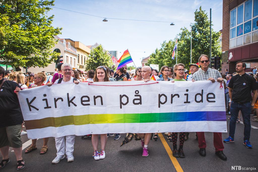 Fem personer bærer et banner med påskriften "Kirken på pride". De går i en parade med mange mennesker. Bak vaier regnbueflagget. Foto.