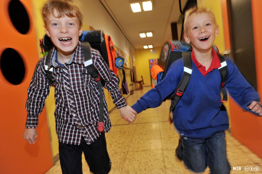 To gutter løper glade hånd i hånd i en korridor med knagger på veggen. Begge har sekk på ryggen og smiler. Foto.