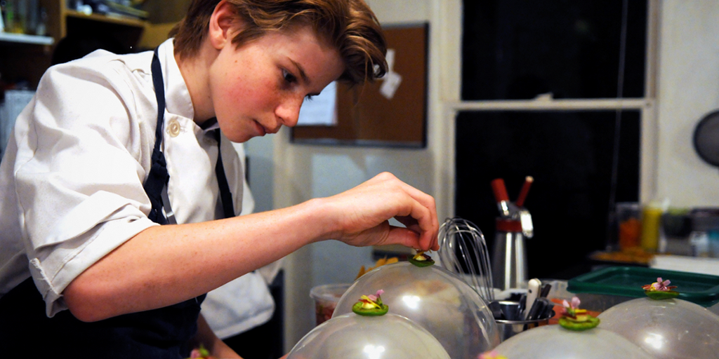 En ung gutt med kokkeklær er på et kjøkken og legger mat på fat. Foto
