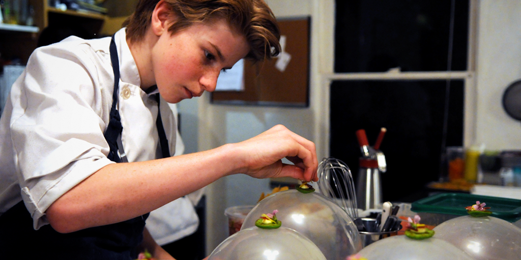 A young boy in chef's clothes is in a kitchen, putting food on plates. Photo.