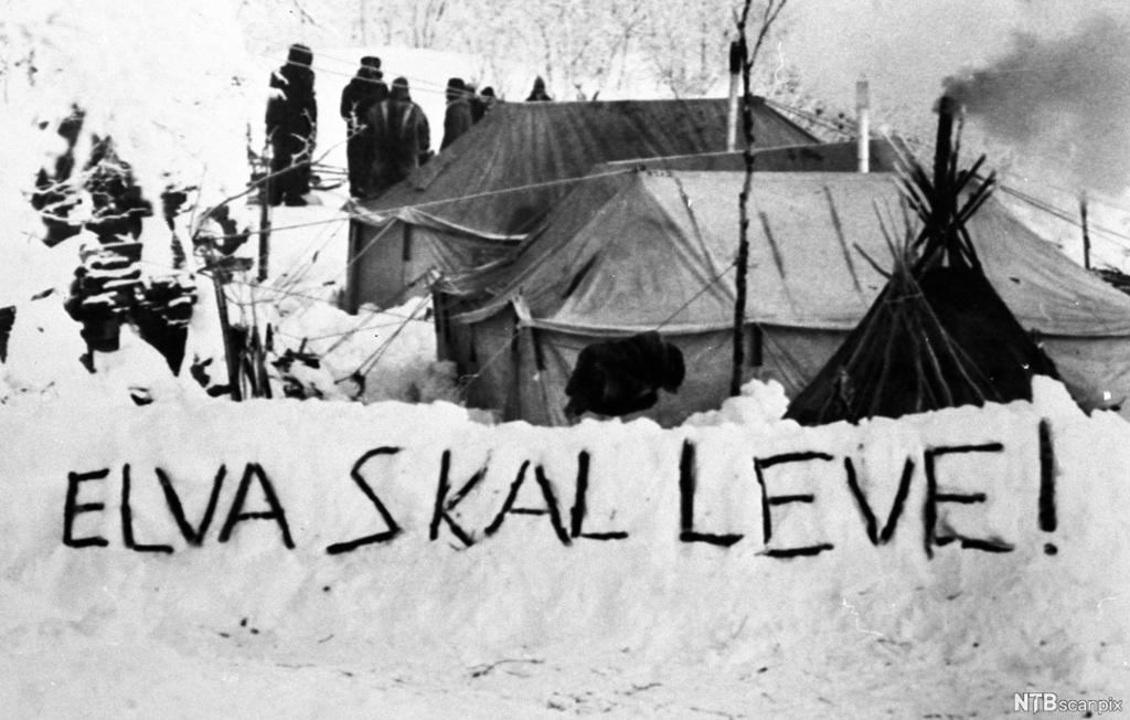 To telt og en lavo er satt opp i snøen. Flere vinterkledde personer står ved teltene. På en snøfonn kan vi se påskriften: "ELVA SKAL LEVE". Svart-hvitt-foto.
