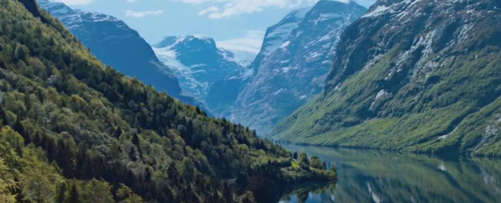 Bratte, grønne fjellsider som speiler seg i en blank fjord. Høye fjell med snø i bakgrunnen. Foto.