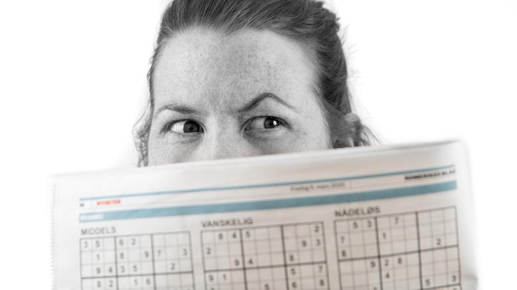 Jente kikkar med skeptisk blikk over ei avis. Foto.