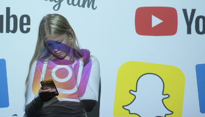 Ei jente ser på mobilen sin. En projektor som viser bilder av logoene til ulike sosiale medier er rettet mot henne. Foto.