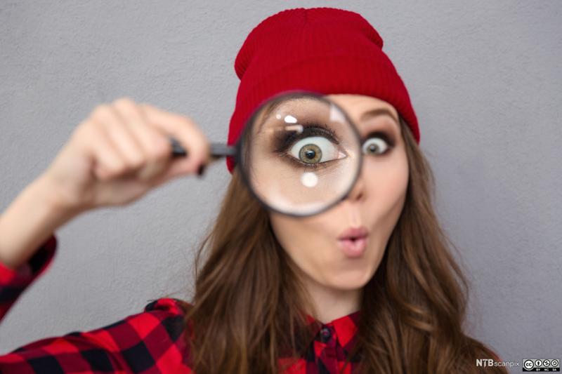 Ung kvinne med rød lue holder et forstørrelsesglass foran det ene øyet. Foto.