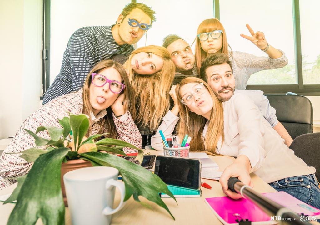 Gruppe med sju unge menneske som tek ein tøysete gruppeselfie ved ein kontorpult. Foto.