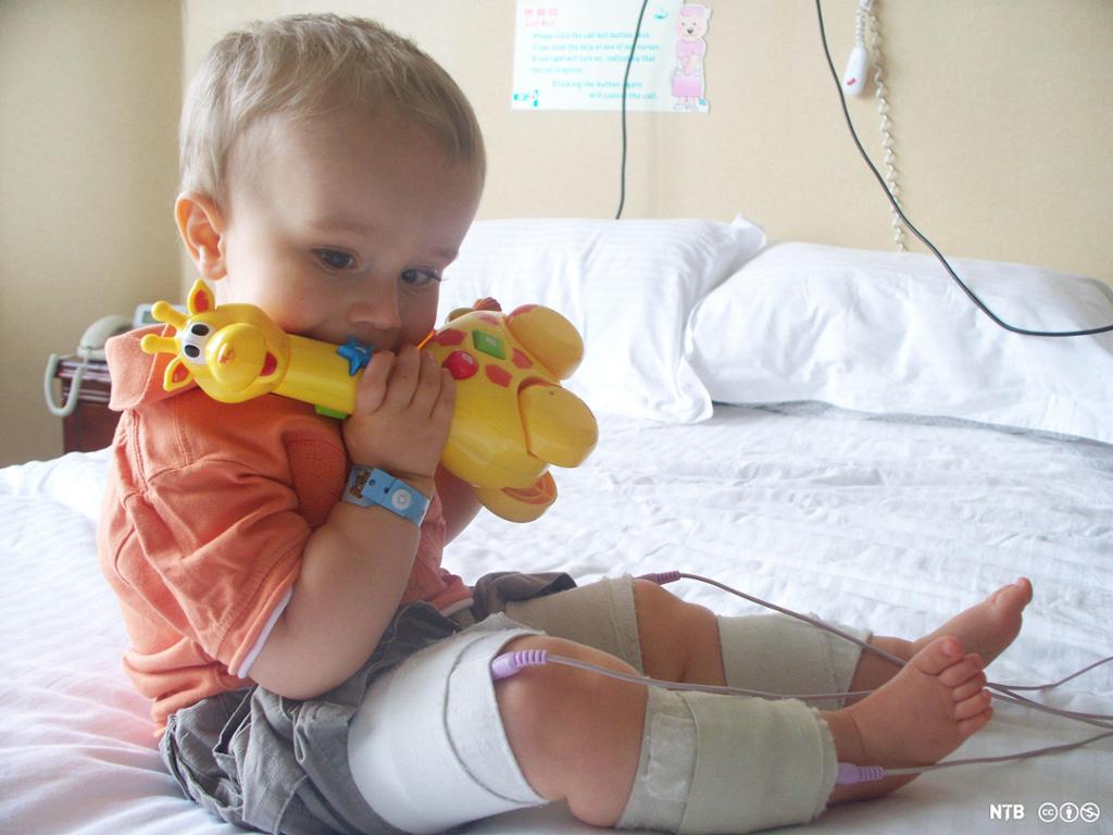 En liten gutt sitter på en seng og biter på en leke mens han får tilført stamceller via en slange. Foto.