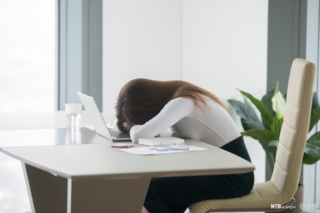 Ung kvinne sitter med hodet i hendene, bøyd over kontorpulten. På pulten står en laptop. Foto.