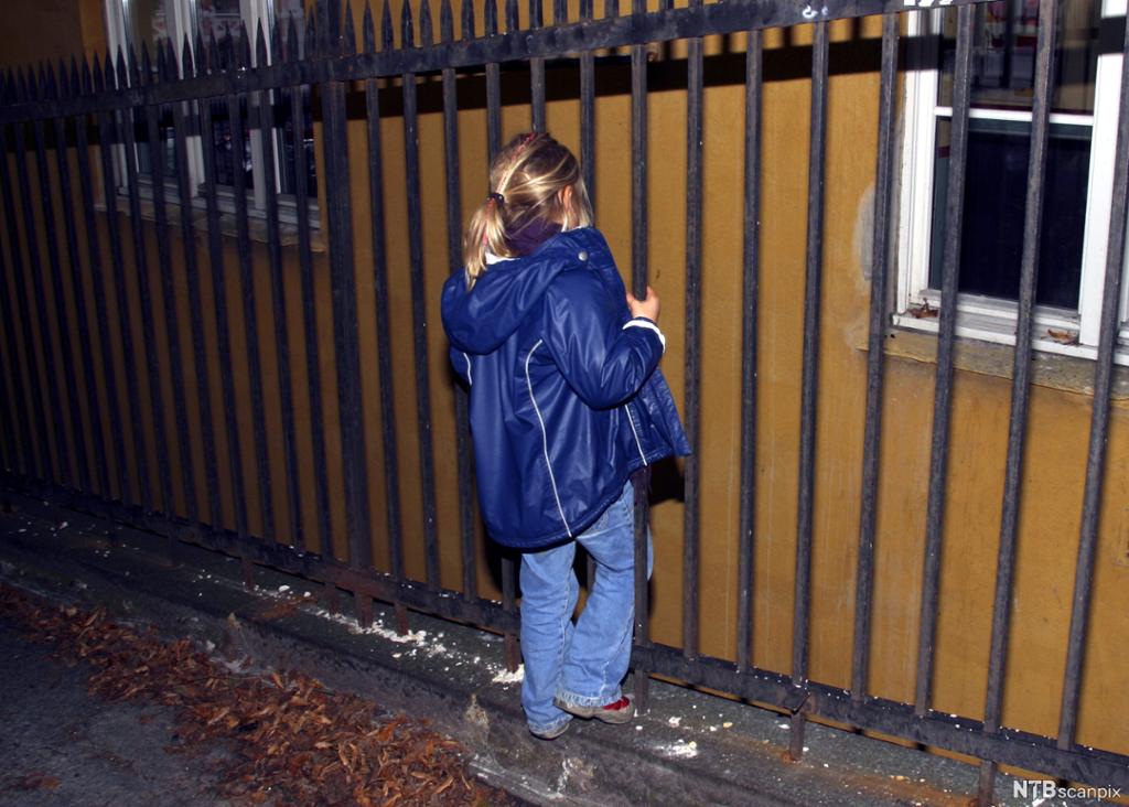 Ei jente står alene og holder seg i et metallgjerde mot en husvegg. Det er mørkt rundt henne, det ligger høstløv på bakken, og hun har åpen jakke. Foto.