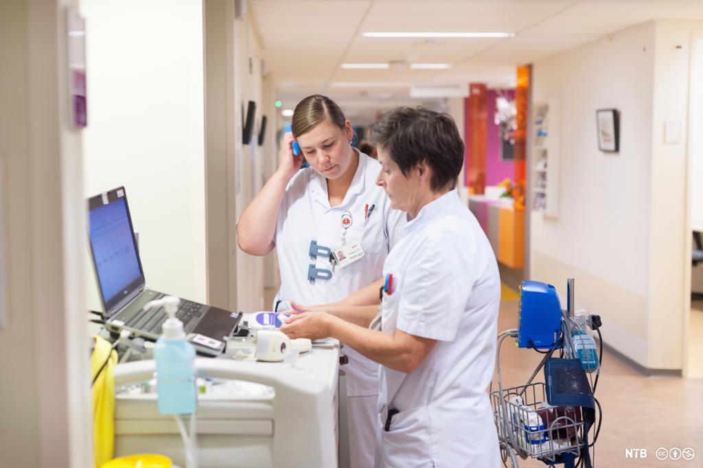 To kvinner med hvite uniformer står ved ei medisintralle i sykehuskorridor. Foto.